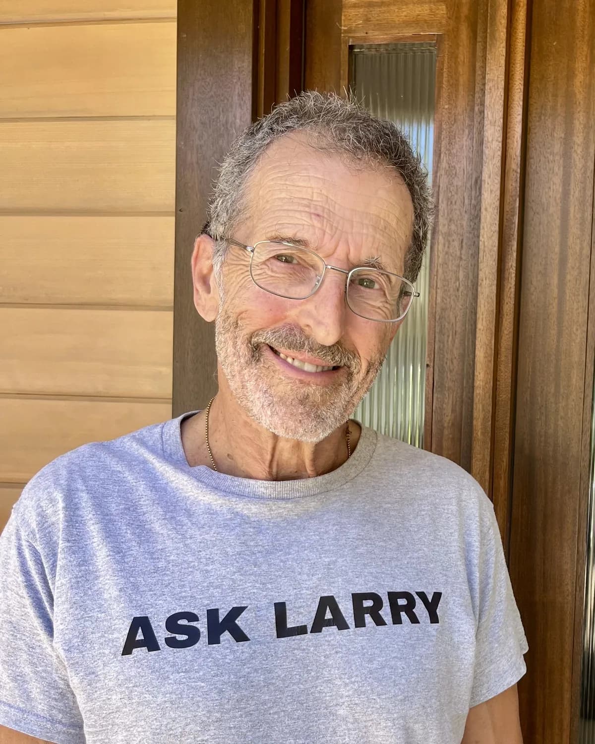 Larry Steingold, smiling, on his front porch in a grey ASK LARRY t-shirt.